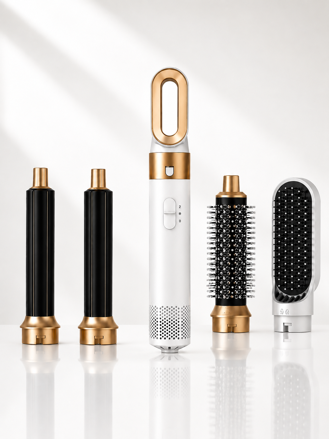 A hair styling tool set displayed on a clean white background with soft shadows and reflections. The set includes a central white and gold wand-style device with three cylindrical attachments in black and gold, and a round brush attachment in black, white, and gold.