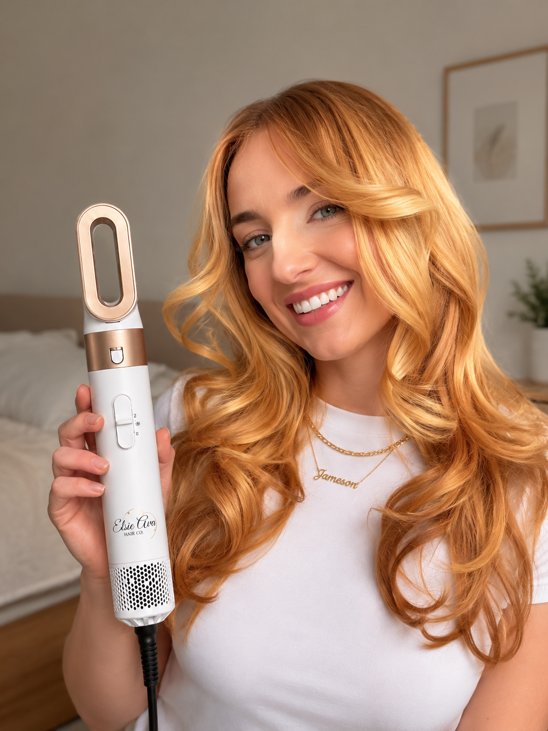 Woman with long blonde hair holding a white and gold hair styling tool in a bedroom setting.
