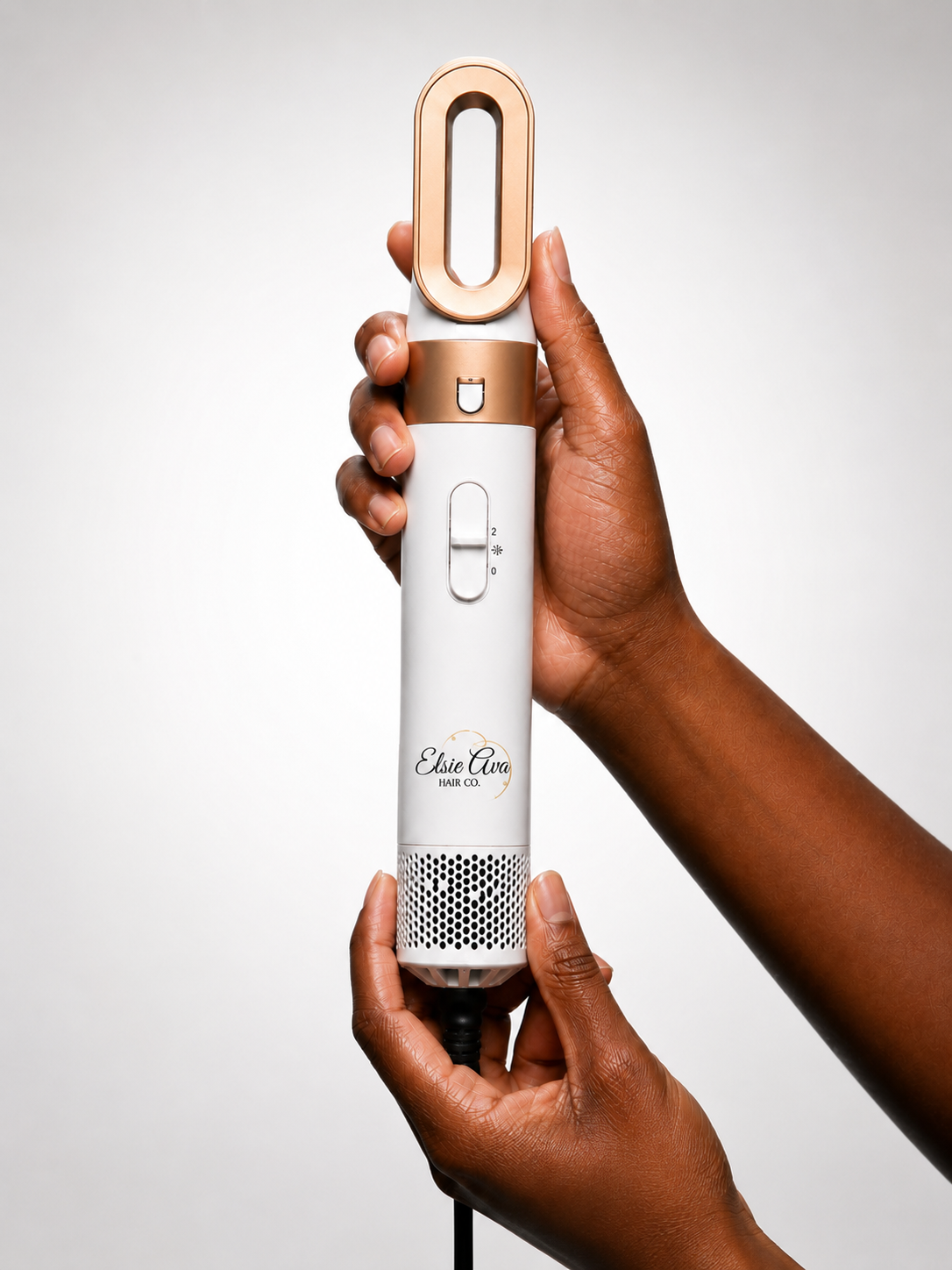 A handheld electric hair dryer held by two hands against a light gray studio background. The dryer has a white body with rose gold accents on the top and around the switch, and a black cord extending from the bottom.